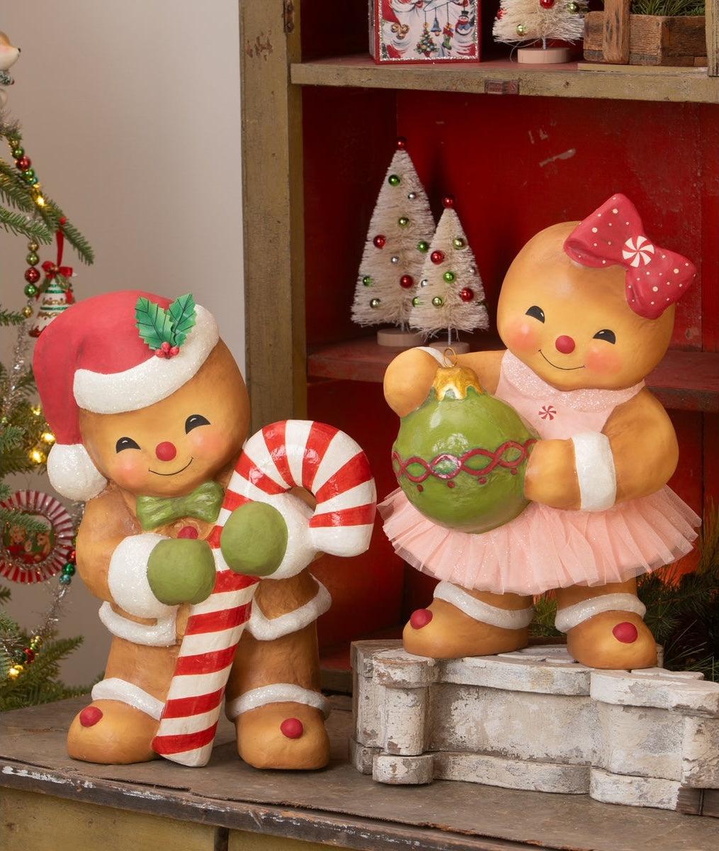 Two gingerbread cookie figurines, one with a candy cane and the other with a green ornament, in a festive setting. Gingerbread Boy George and Gingerbread Girl Joy.