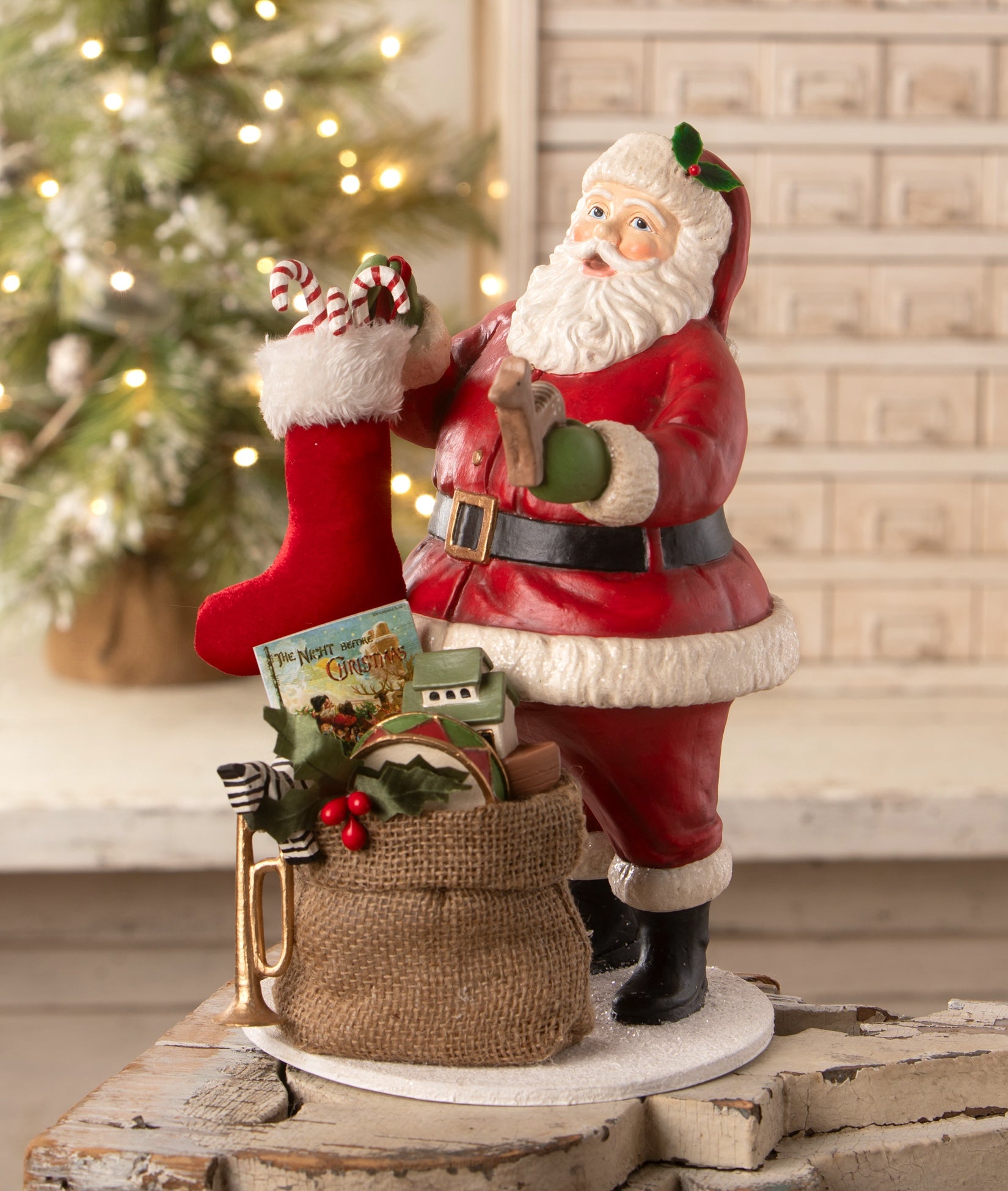 Decorative Santa Claus figure with a stocking and gift bag in front of a Christmas tree. Santa Filling the Stocking by Bethany Lowe.
